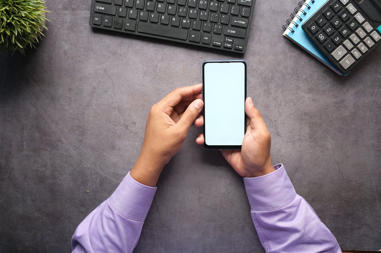 why-choose-us Man in purple shirt holding a smartphone with blank screen at office desk with keyboard, notebook, and calculator.